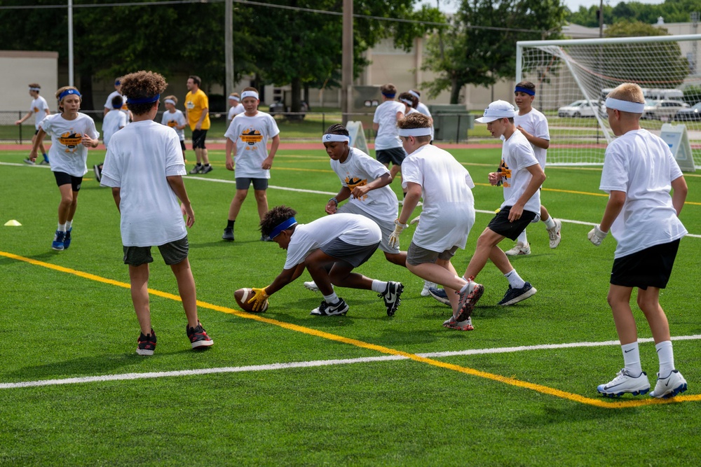 A group of youth players participate in an outdoor football camp or drill session on a green artificial turf field. One player is bent down picking up or fumbling a football while others converge around the ball. The participants are wearing matching white t-shirts with a logo and colorful headbands or visors, suggesting an organized sports camp.