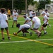 A group of youth players participate in an outdoor football camp or drill session on a green artificial turf field. One player is bent down picking up or fumbling a football while others converge around the ball. The participants are wearing matching white t-shirts with a logo and colorful headbands or visors, suggesting an organized sports camp.