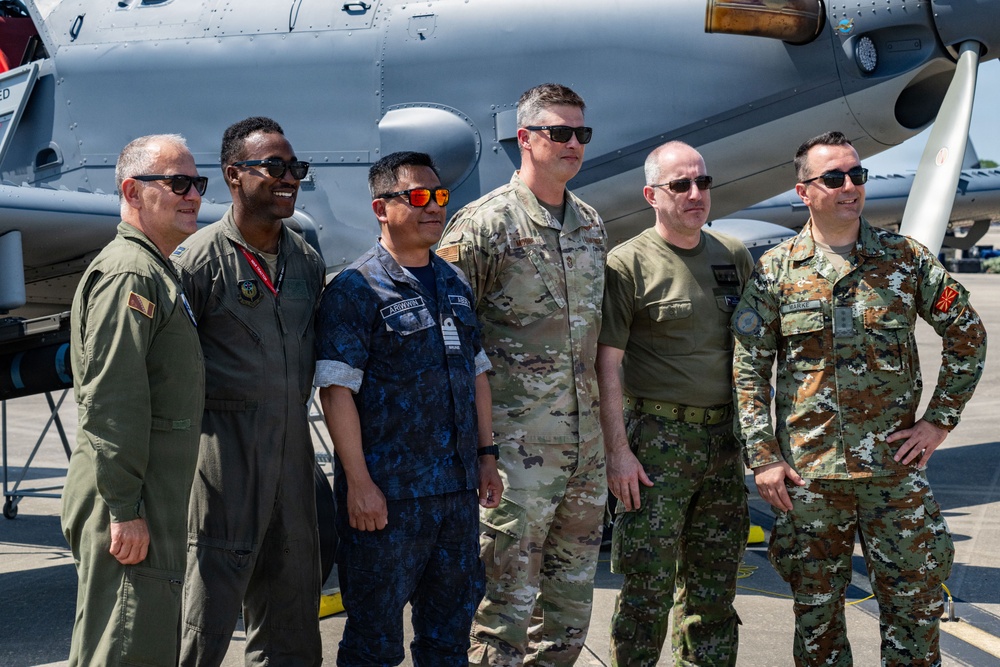 Six military personnel from what appear to be multiple nations are posing together in front of a military aircraft on a flight line or tarmac. The group displays a variety of uniforms and camouflage patterns, suggesting a multinational military gathering or exercise. The setting and attire indicate a formal or semi-formal group photo opportunity at an airbase.
