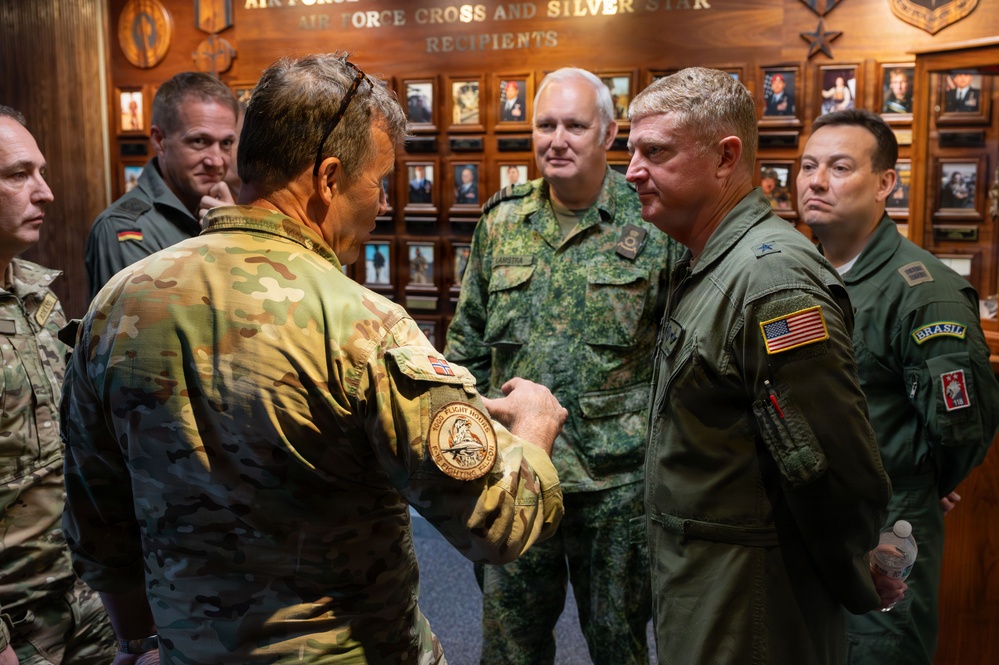 A group of multinational military officers are gathered in what appears to be a hall of honor displaying 'Air Force Cross and Silver Star Recipients' plaques and portraits. Several officers in different national uniforms are engaged in conversation, with one officer appearing to show or discuss a patch on his sleeve. The group represents multiple nations including the United States, Germany, Brazil, and at least one other country.