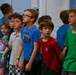A group of approximately 8-10 children, appearing to be elementary school age, are standing together in a line against a mirrored wall, looking upward or forward attentively. The setting appears to be an indoor facility such as a school, gym, or community center. The children are dressed casually in colorful t-shirts and appear engaged in a shared activity or presentation.