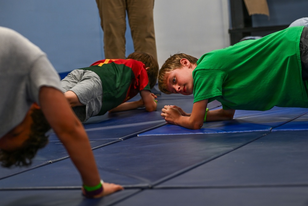 Several children are performing plank or push-up exercises on blue gymnastics mats in what appears to be a gym or fitness facility. A boy in a green t-shirt is looking directly at the camera while holding a plank position. An adult's legs are visible in the background, suggesting supervision.
