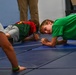 Several children are performing plank or push-up exercises on blue gymnastics mats in what appears to be a gym or fitness facility. A boy in a green t-shirt is looking directly at the camera while holding a plank position. An adult's legs are visible in the background, suggesting supervision.