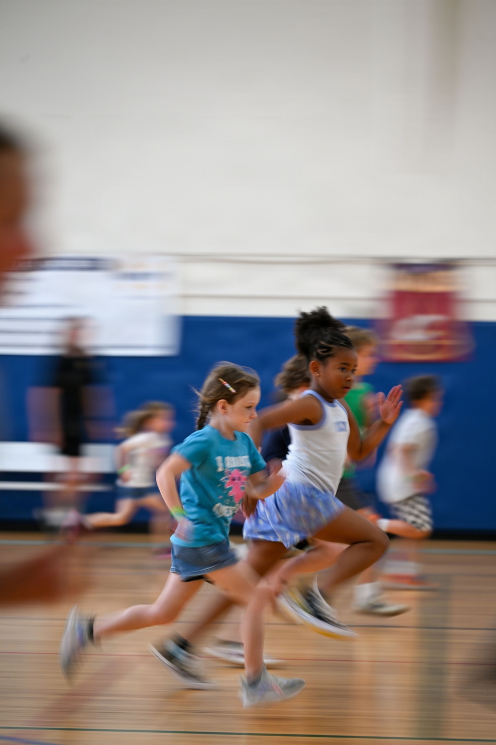 A group of young children are running at full speed across an indoor gymnasium floor. The image captures significant motion blur, conveying the energy and speed of the activity. Two girls are prominently featured in the foreground — one in a teal t-shirt and one in a white tank top with a blue skirt.