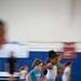 A group of young children are running at full speed across an indoor gymnasium floor. The image captures significant motion blur, conveying the energy and speed of the activity. Two girls are prominently featured in the foreground — one in a teal t-shirt and one in a white tank top with a blue skirt.