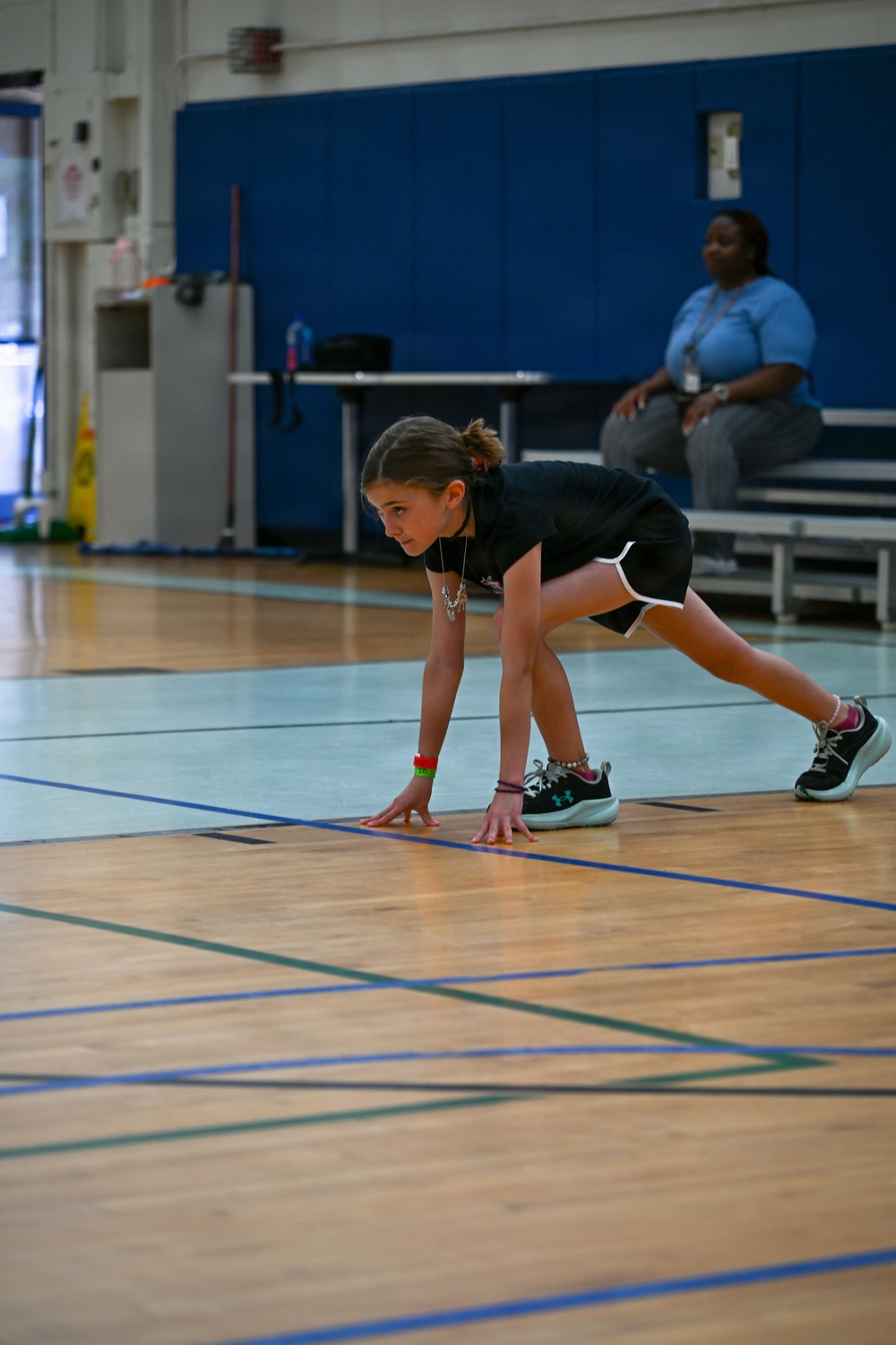 A young girl is in a crouched, ready position on a gymnasium floor, appearing to be at the start of a race or drill. She is wearing a black athletic outfit with Under Armour sneakers and various bracelets and a necklace. An adult supervisor is visible in the background seated on bleachers.