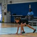 A young girl is in a crouched, ready position on a gymnasium floor, appearing to be at the start of a race or drill. She is wearing a black athletic outfit with Under Armour sneakers and various bracelets and a necklace. An adult supervisor is visible in the background seated on bleachers.