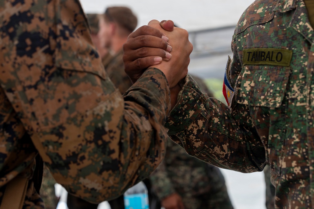 Two military personnel from different nations are clasping hands in a firm handshake or greeting gesture. One soldier wears US Marine Corps MARPAT woodland camouflage, while the other wears a different digital camouflage pattern with a name tape reading 'TAMBALO' and what appears to be a Philippine flag patch. The image likely depicts a bilateral military exercise or cooperative engagement between US and Philippine armed forces.