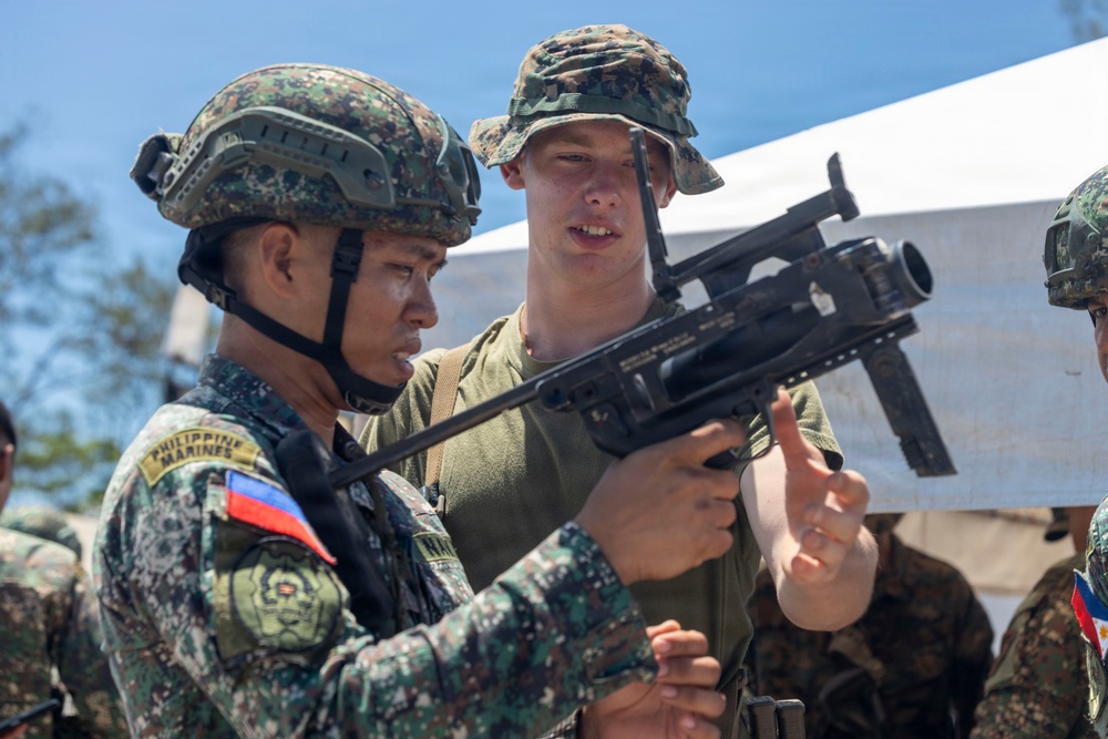 The image shows a joint military training exercise between Philippine Marines and what appears to be a U.S. Marine. A Philippine Marine wearing a helmet and camouflage uniform with a 'Philippine Marines' patch is handling a weapon, while a taller soldier in a boonie hat and olive t-shirt observes and assists. The setting appears to be an outdoor training area with a tent canopy visible in the background.