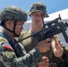 The image shows a joint military training exercise between Philippine Marines and what appears to be a U.S. Marine. A Philippine Marine wearing a helmet and camouflage uniform with a 'Philippine Marines' patch is handling a weapon, while a taller soldier in a boonie hat and olive t-shirt observes and assists. The setting appears to be an outdoor training area with a tent canopy visible in the background.