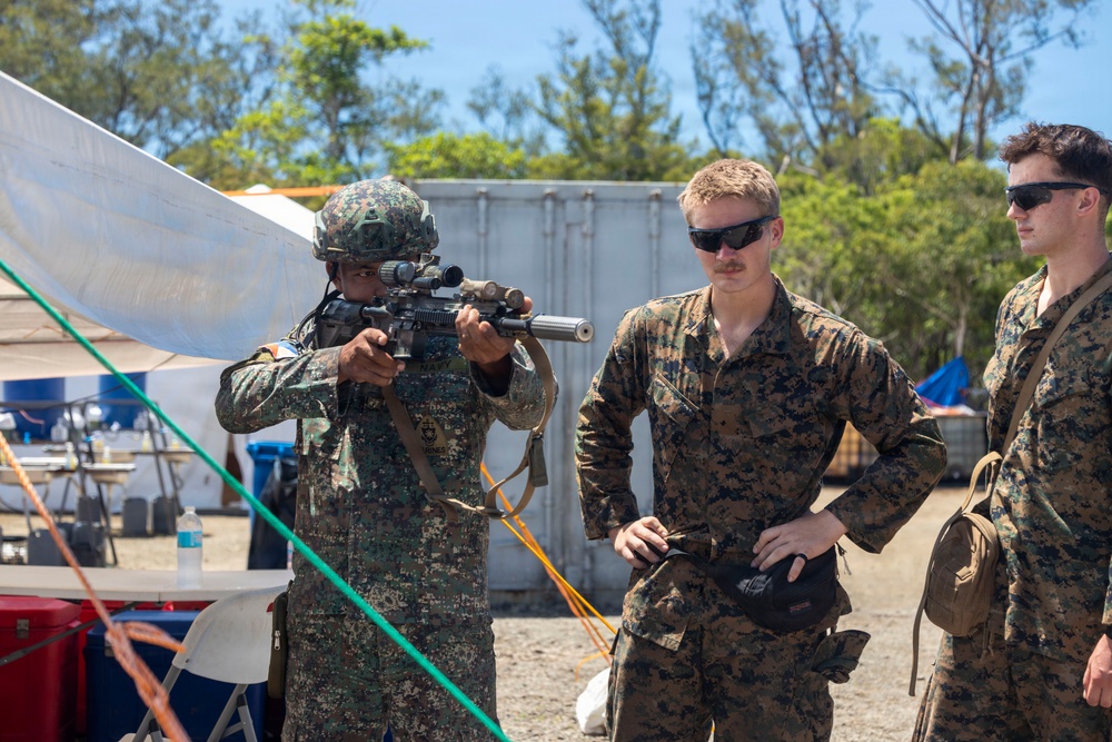 Three military personnel are gathered in an outdoor field environment, with one soldier in a helmet and different camouflage pattern aiming a rifle equipped with a scope and suppressor. Two other personnel in matching digital camouflage uniforms observe, suggesting a weapons demonstration or training exchange. The setting appears to be a temporary military camp or exercise area.
