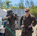 Three military personnel are gathered in an outdoor field environment, with one soldier in a helmet and different camouflage pattern aiming a rifle equipped with a scope and suppressor. Two other personnel in matching digital camouflage uniforms observe, suggesting a weapons demonstration or training exchange. The setting appears to be a temporary military camp or exercise area.