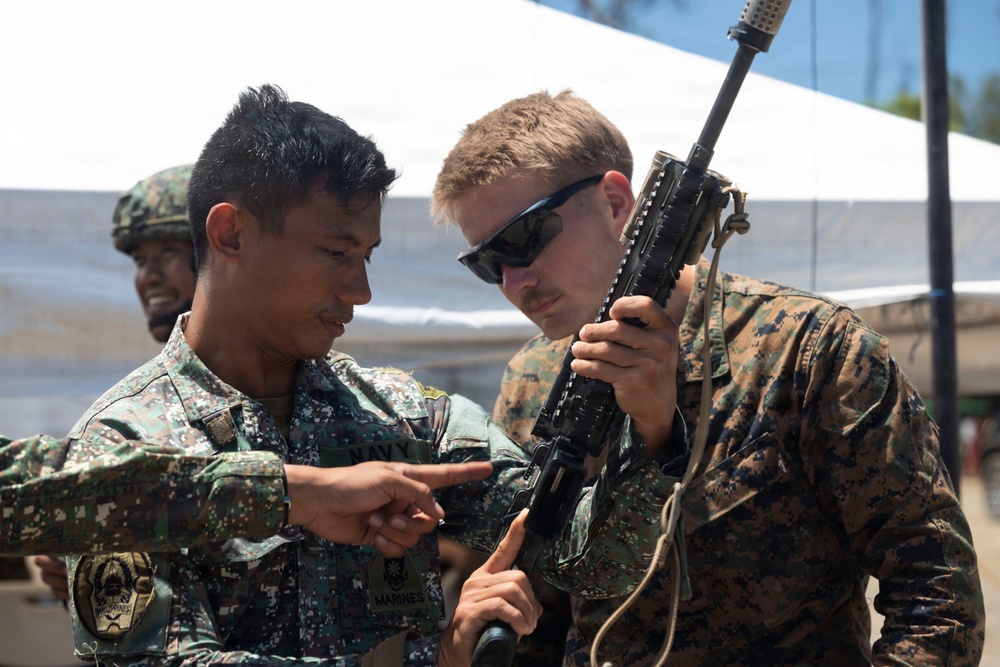 Two military personnel are examining a rifle together in what appears to be a joint training exercise. One individual wears a Philippine Navy/Marines uniform and is pointing at a component of the weapon, while the other wears a US Marine Corps MARPAT woodland uniform and is holding the rifle upright for inspection. A third uniformed individual is visible in the background near a white tent structure.