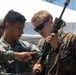 Two military personnel are examining a rifle together in what appears to be a joint training exercise. One individual wears a Philippine Navy/Marines uniform and is pointing at a component of the weapon, while the other wears a US Marine Corps MARPAT woodland uniform and is holding the rifle upright for inspection. A third uniformed individual is visible in the background near a white tent structure.