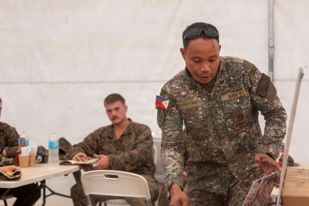 A Philippine Navy servicemember named Tamayao, wearing digital camouflage uniform with a Philippine flag patch, is leaning over a table and reviewing printed materials inside a military tent. In the background, at least two US Marines in MARPAT uniforms are seated at folding tables, appearing to take notes. The setting suggests a joint military briefing or training exercise.