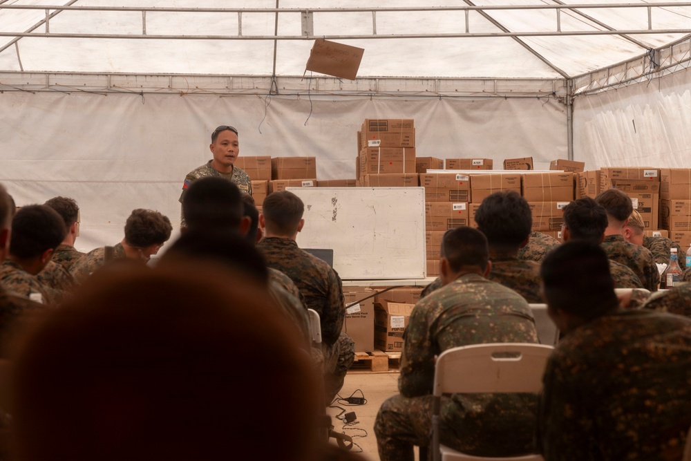 A military briefing or instruction session is taking place inside a large tent or temporary structure. One soldier stands at the front addressing a seated audience of uniformed personnel, with a whiteboard and stacked cardboard boxes visible behind him. The attendees appear to wear two different camouflage uniform patterns, suggesting a multinational or joint military exercise.