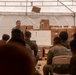 A military briefing or instruction session is taking place inside a large tent or temporary structure. One soldier stands at the front addressing a seated audience of uniformed personnel, with a whiteboard and stacked cardboard boxes visible behind him. The attendees appear to wear two different camouflage uniform patterns, suggesting a multinational or joint military exercise.