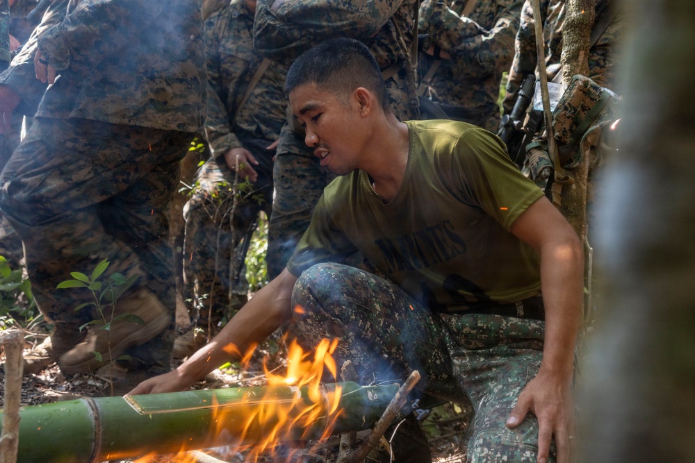 A soldier in a green t-shirt and camouflage pants is blowing on a fire built around a green bamboo segment in a jungle or forested environment. He is surrounded by other personnel wearing woodland digital camouflage uniforms, suggesting a military survival training exercise. The scene depicts a fire-starting or jungle survival skills demonstration.