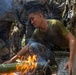 A soldier in a green t-shirt and camouflage pants is blowing on a fire built around a green bamboo segment in a jungle or forested environment. He is surrounded by other personnel wearing woodland digital camouflage uniforms, suggesting a military survival training exercise. The scene depicts a fire-starting or jungle survival skills demonstration.