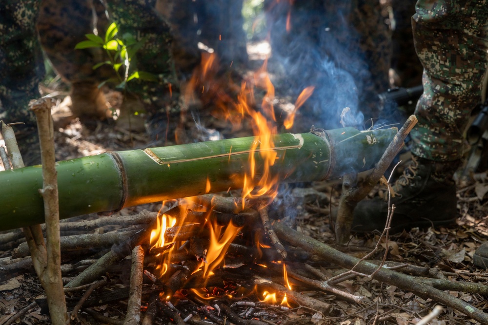The image shows a large green bamboo stalk being cooked over an open fire in a forest setting. The bamboo appears to have been split or scored along its length and is elevated on sticks above burning wood and embers. Individuals wearing camouflage military-style uniforms are visible in the background, suggesting a survival training or field cooking exercise.