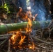 The image shows a large green bamboo stalk being cooked over an open fire in a forest setting. The bamboo appears to have been split or scored along its length and is elevated on sticks above burning wood and embers. Individuals wearing camouflage military-style uniforms are visible in the background, suggesting a survival training or field cooking exercise.