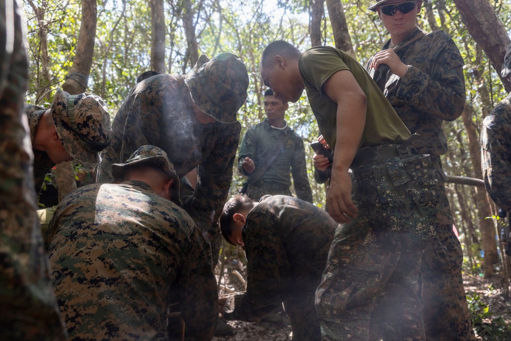 A group of military personnel in camouflage uniforms are gathered in a forested area, focused on a ground-level task with visible smoke present. The group appears to include members wearing at least two different camouflage patterns, suggesting a joint or multinational training exercise. The activity appears to be a field training exercise, possibly involving fire-starting or survival skills.