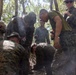 A group of military personnel in camouflage uniforms are gathered in a forested area, focused on a ground-level task with visible smoke present. The group appears to include members wearing at least two different camouflage patterns, suggesting a joint or multinational training exercise. The activity appears to be a field training exercise, possibly involving fire-starting or survival skills.