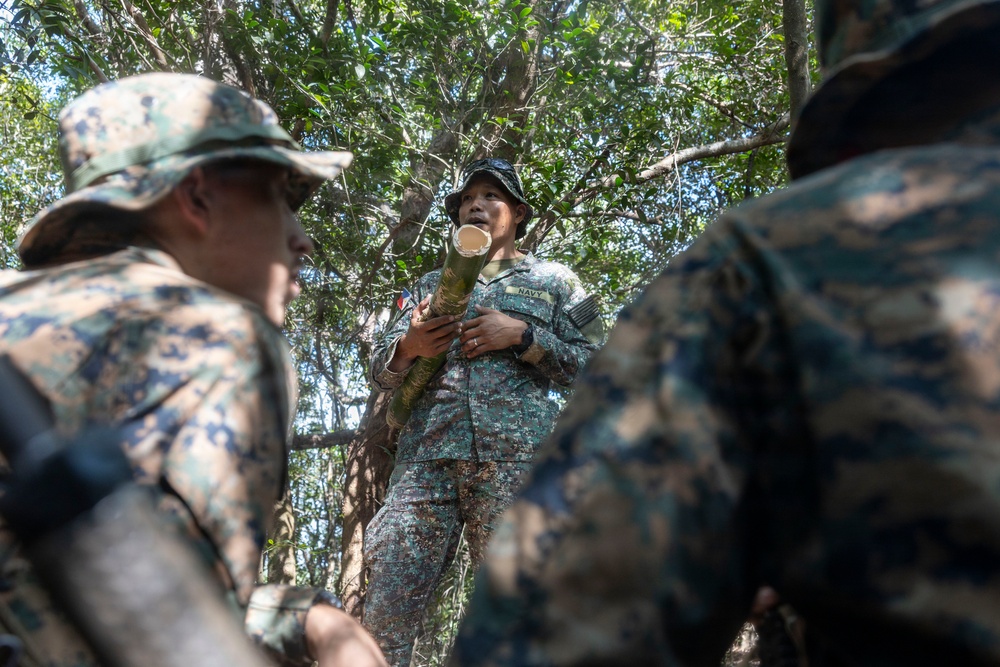 A military training or instruction session taking place in a forested outdoor environment. A central figure wearing a 'NAVY' labeled camouflage uniform is holding a section of bamboo and appears to be instructing or demonstrating something to other personnel. The surrounding individuals are wearing MARPAT (Marine pattern) camouflage uniforms, suggesting a joint or multinational military exercise.