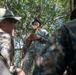 A military training or instruction session taking place in a forested outdoor environment. A central figure wearing a 'NAVY' labeled camouflage uniform is holding a section of bamboo and appears to be instructing or demonstrating something to other personnel. The surrounding individuals are wearing MARPAT (Marine pattern) camouflage uniforms, suggesting a joint or multinational military exercise.