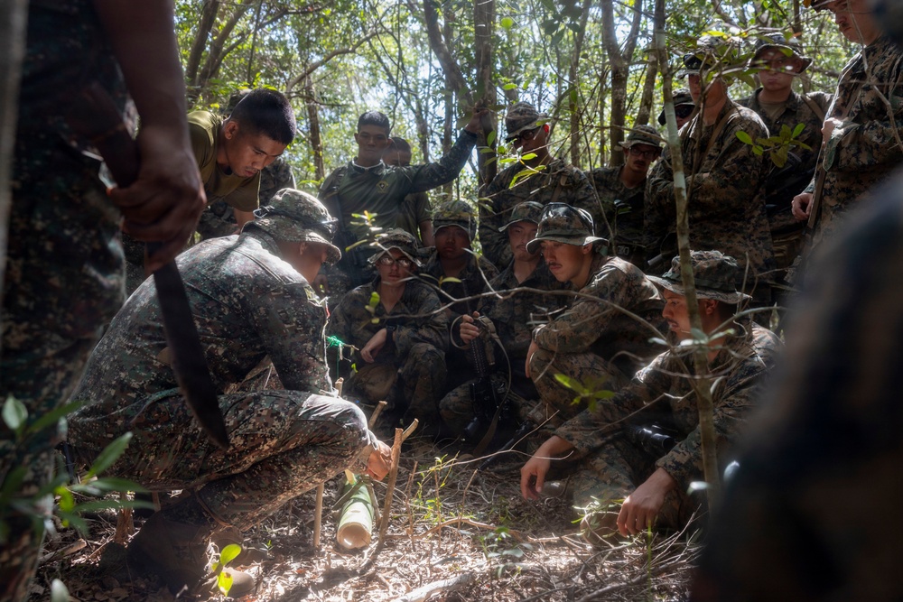 A group of military personnel in camouflage uniforms are gathered in a forested area, observing a demonstration or instruction being conducted on the ground. The scene appears to be a jungle survival or field craft training exercise, with one individual demonstrating a technique using bamboo or wooden materials. The mixed uniforms suggest a multinational or joint military training exercise.