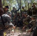 A group of military personnel in camouflage uniforms are gathered in a forested area, observing a demonstration or instruction being conducted on the ground. The scene appears to be a jungle survival or field craft training exercise, with one individual demonstrating a technique using bamboo or wooden materials. The mixed uniforms suggest a multinational or joint military training exercise.