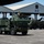 The image shows multiple olive-drab military vehicles, consistent with M142 HIMARS or similar rocket artillery systems, positioned on a paved military installation apron. At least two soldiers are visible atop the vehicles, and a large open-sided hangar marked 'H3' is visible in the background. The setting appears to be a military airfield or port facility.