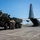 A U.S. military scene on an airfield tarmac showing a HIMARS (High Mobility Artillery Rocket System) vehicle positioned adjacent to a C-130 Hercules transport aircraft. Several soldiers in U.S. Army combat uniforms are gathered between the two vehicles, apparently coordinating a loading or offloading operation. The aircraft bears tail code 'YJ' and serial number 37406, indicating assignment to Yokota Air Base, Japan.