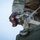 A person wearing military multicam/OCP camouflage uniform is manipulating a metal ratchet strap buckle or tie-down mechanism on what appears to be a military green equipment case or container. The low-angle shot provides a close-up view of the hands working with the securing hardware. The image captures a logistics or equipment-securing task in a military context.