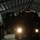 A large armored tactical vehicle is positioned inside a dimly lit industrial hangar or maintenance bay with its headlights illuminated. Several personnel in military or law enforcement uniforms are present, with two individuals standing to the left and at least two others on or near the vehicle. The scene appears to depict a briefing, inspection, or staging operation in a low-light environment.