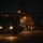 A nighttime military logistics operation at an airfield showing a large military vehicle being loaded or unloaded from a transport aircraft. The scene is illuminated primarily by the vehicle's headlights and the aircraft's interior lighting. Several personnel in military and civilian attire are visible near the aircraft's open rear cargo ramp.