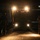A nighttime military or aviation logistics operation showing a large vehicle being guided into or out of what appears to be a military transport aircraft. Two personnel in uniform are visible on either side of the vehicle, with one appearing to give hand signals to direct the driver. The scene is illuminated primarily by the vehicle's headlights and work lights, with a partially visible 'SIA Engineering' sign in the background.