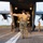 A military loadmaster in multicam camouflage uniform is marshaling a military vehicle (appears to be an LMTV or similar tactical truck) into the rear cargo ramp of a C-130 Hercules transport aircraft on a tarmac. The loadmaster is using hand signals with arms outstretched while wearing communications headset. Two additional personnel are visible — one crouching near the ramp and one standing to the right observing.