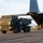 A military HIMARS (High Mobility Artillery Rocket System) vehicle is being loaded onto or unloaded from a U.S. Air Force C-130 Hercules transport aircraft on a tarmac. The aircraft tail markings show 'YJ' and serial number '37406' with unit designation '810', indicating it belongs to the 374th Airlift Wing based at Yokota Air Base, Japan. Personnel in military uniforms are visible near the aircraft's rear cargo ramp.