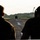 Two individuals in military-style attire are viewed from behind, observing a runway or airstrip where a military vehicle is visible in the distance. The scene appears to take place at dawn or dusk based on the soft, hazy lighting. A treeline is visible in the background, suggesting a tropical or subtropical environment.