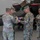 Task Force Vipers transfer authority to Task Force Gunfighters A group of U.S. Army soldiers in OCP (Operational Camouflage Pattern) uniforms are performing a military flag ceremony, likely a casing or uncasing of colors, inside an aircraft hangar. Several soldiers are handling what appears to be a folded guidon or unit flag with blue and gold/orange colors. An Apache helicopter (AH-64) is visible in the background.