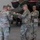 Task Force Vipers transfer authority to Task Force Gunfighters Four U.S. Army soldiers in OCP (Operational Camouflage Pattern) uniforms are gathered on a flight line or hangar apron, handling what appears to be a green fabric item, possibly a flag or cover. A military helicopter, likely an AH-64 Apache, is visible in the background. The setting appears to be an outdoor or semi-covered military aviation facility.