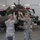 Task Force Vipers transfer authority to Task Force Gunfighters Several U.S. Army soldiers in OCP (Operational Camouflage Pattern) uniforms are gathered inside a military hangar, appearing to engage in a celebratory high-five or clapping gesture. In the background, a military attack helicopter (consistent with an AH-64 Apache) is visible with panels open, suggesting maintenance activity. A green cloth or item is visible mid-air near the soldiers.