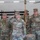 Task Force Vipers transfer authority to Task Force Gunfighters Four U.S. Army soldiers in Operational Camouflage Pattern (OCP) uniforms are participating in a formal military ceremony inside what appears to be a hangar or maintenance facility. The central figure is rendering a salute, while the others stand at attention. A soldier in the background is holding a guidon or unit flag on a wooden staff.