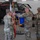 Task Force Vipers transfer authority to Task Force Gunfighters A military ceremony is taking place inside an aircraft hangar, with soldiers in U.S. Army OCP (Operational Camouflage Pattern) uniforms unfurling or casing a unit guidon/colors. The blue flag with gold fringe and accompanying streamer ribbons suggest a formal activation, deactivation, or change of command ceremony. An AH-64 Apache attack helicopter is visible in the background.