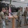 Task Force Vipers transfer authority to Task Force Gunfighters A military ceremony is taking place inside an aircraft hangar, with a soldier speaking at a wooden podium equipped with a microphone. Several soldiers stand at attention in the background holding flags, including the American flag and what appears to be a unit guidon. Apache attack helicopters are visible in the background, suggesting this is an aviation unit ceremony.