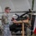 Task Force Vipers transfer authority to Task Force Gunfighters A U.S. Army officer in OCP (Operational Camouflage Pattern) uniform speaks at a wooden podium during a formal military ceremony held inside an aircraft hangar. Behind him, a soldier holds multiple flags including the U.S. flag and what appears to be the Polish national flag, suggesting a joint or NATO-related event. An Apache attack helicopter (likely AH-64) is visible in the background.