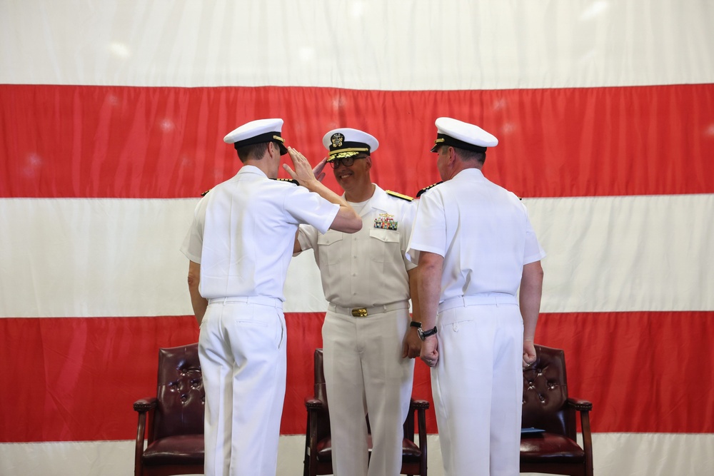 Three U.S. Navy officers in white dress uniforms are participating in a formal military ceremony, likely a change of command, in front of a large American flag backdrop. The central officer, who displays admiral rank insignia and a medal ribbon rack, is receiving a salute from the officer on the left. Two dark wooden chairs are positioned behind the officers on either side.