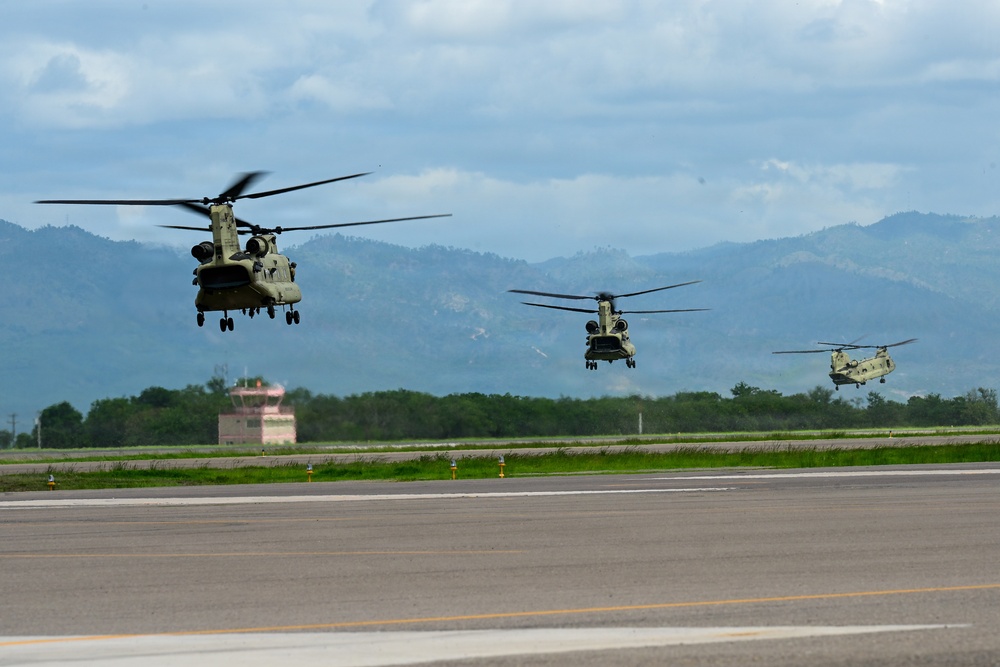 Three military tandem-rotor helicopters, identifiable as Boeing CH-47 Chinooks, are captured in low-level flight over an airfield tarmac in a formation sequence. The helicopters appear to be either taking off or landing in succession along the runway. A mountainous, green landscape and an air traffic control tower are visible in the background under overcast skies.