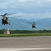 Three military tandem-rotor helicopters, identifiable as Boeing CH-47 Chinooks, are captured in low-level flight over an airfield tarmac in a formation sequence. The helicopters appear to be either taking off or landing in succession along the runway. A mountainous, green landscape and an air traffic control tower are visible in the background under overcast skies.